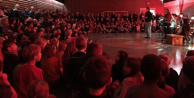 Auf dem Bild sind Lehrer, Eltern und Sch&uuml;ler_innen beim Weihnachtssingen in der Sporthalle zu sehen. Ein paar Kinder spielen dabei Instrumente.
