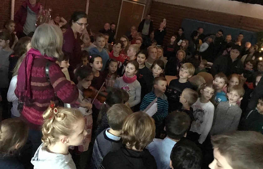 Auf dem Bild sind viele Kinder, Eltern und Lehrer beim Singen in der Turnhalle zu sehen. Zudem spielt ein Kind Geige.