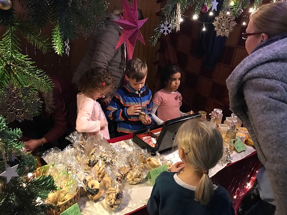Auf dem Bild sind ein paar Kinder und Eltern an ihrem Stand auf dem Wintermarkt zu sehen. Der Stand ist weihnachtlich geschmückt und sie scheinen Spaß am Verkaufen ihrer selbstgebastelten Artikeln zu haben.